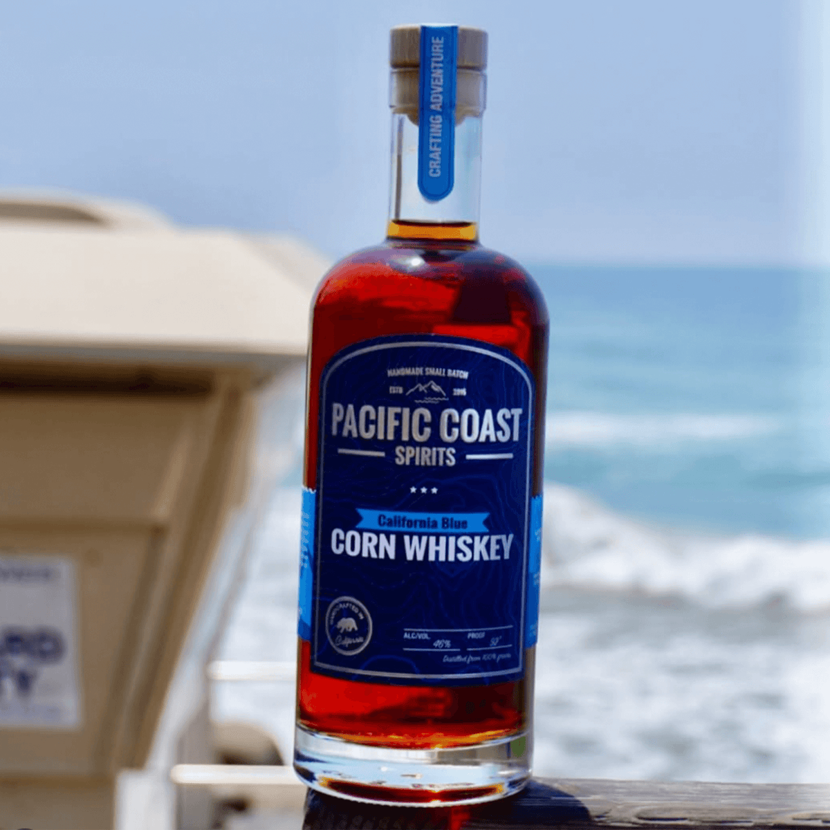 Bottle of Pacific Coast California Blue Corn Whiskey against ocean backdrop, showcasing handcrafted adventure spirit.
