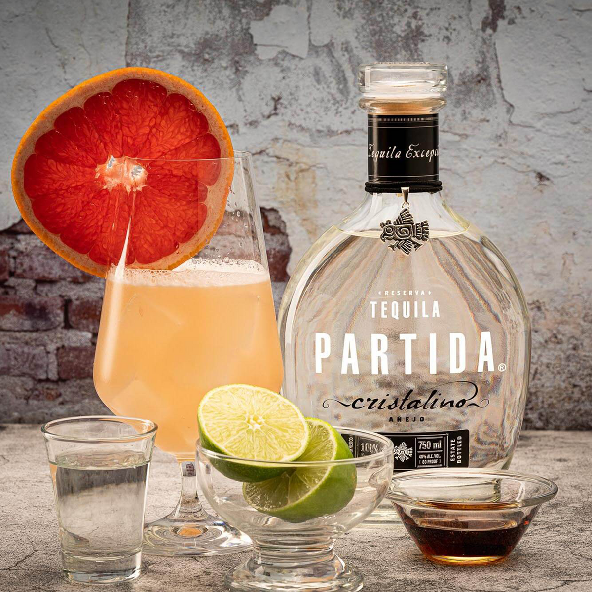 Partida Cristalino Anejo Tequila bottle with cocktails and citrus garnishes on a rustic background.