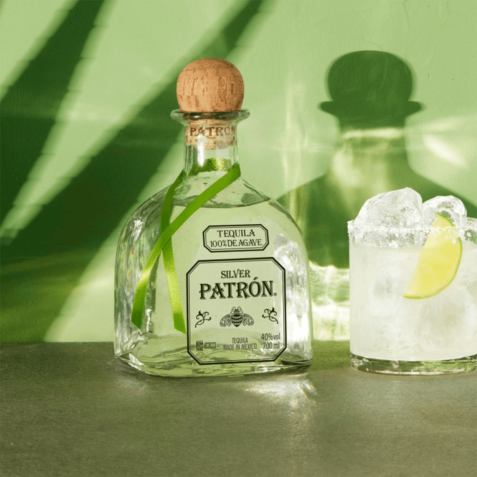 Patrón Silver Tequila bottle with lime garnish next to a cocktail glass on a green background.