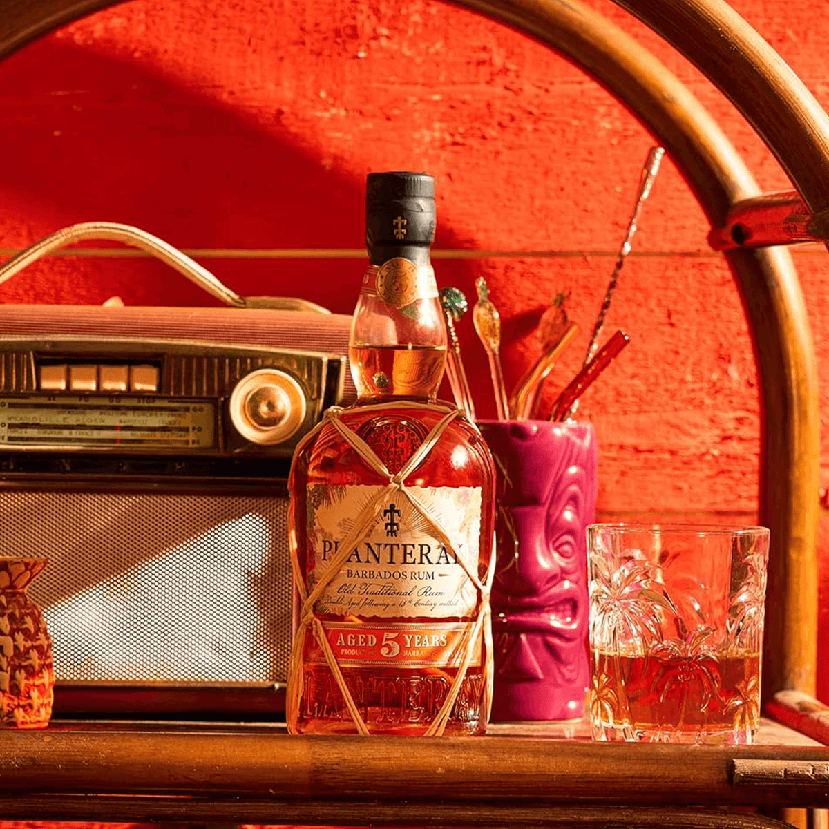 A bottle of Planteray Barbados 5 Year Rum displayed next to a vintage radio and glassware against a vibrant red background.