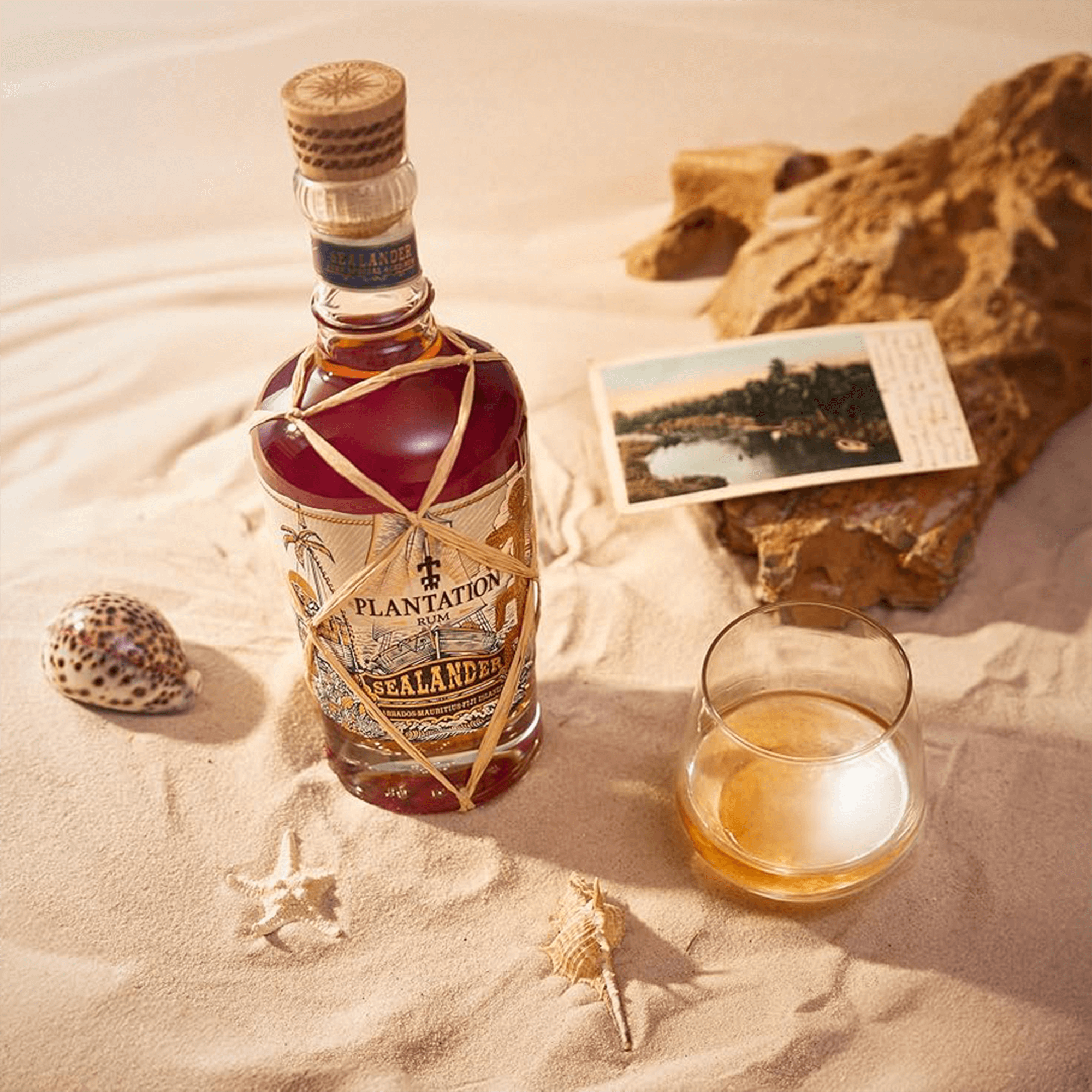 Planteray Sealander Rum bottle with a glass, seashells, and vintage postcard on sandy surface.