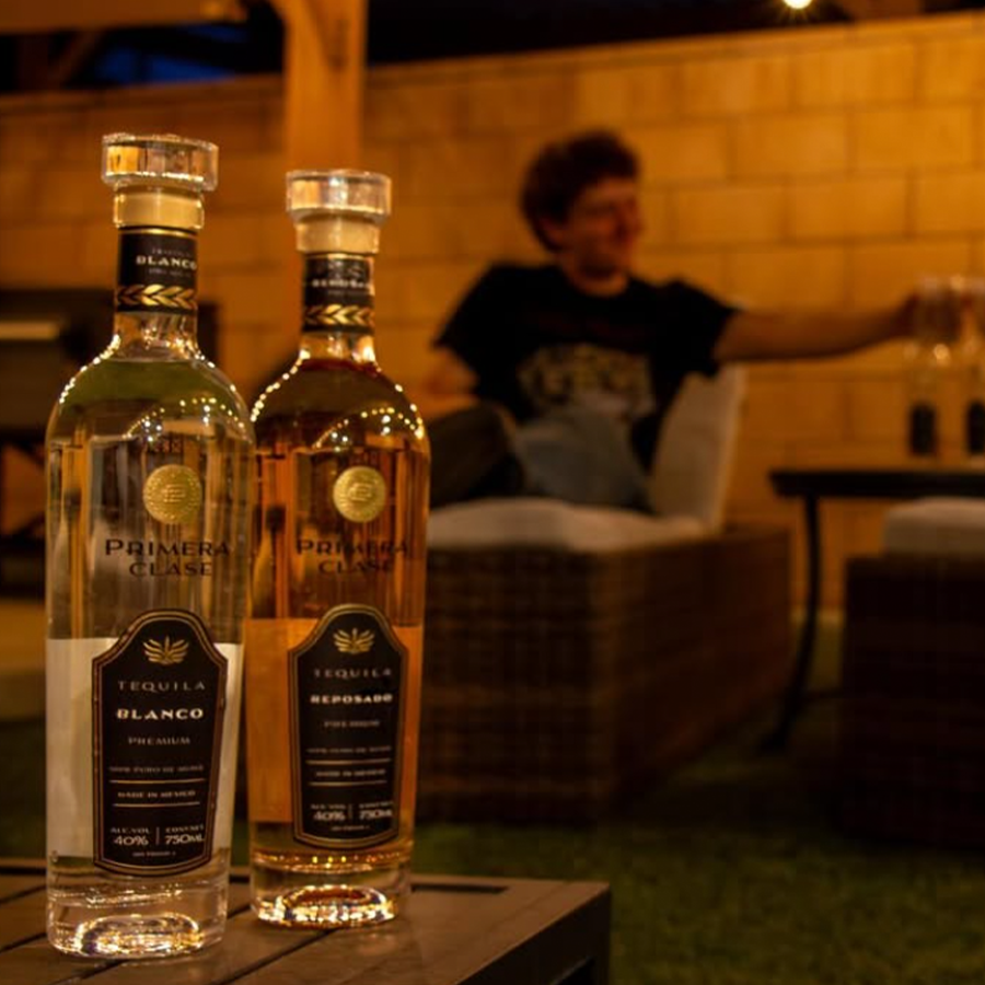 Two bottles of Primera Clase tequila, Blanco and Reposado, showcased in an outdoor setting with a person in the background.