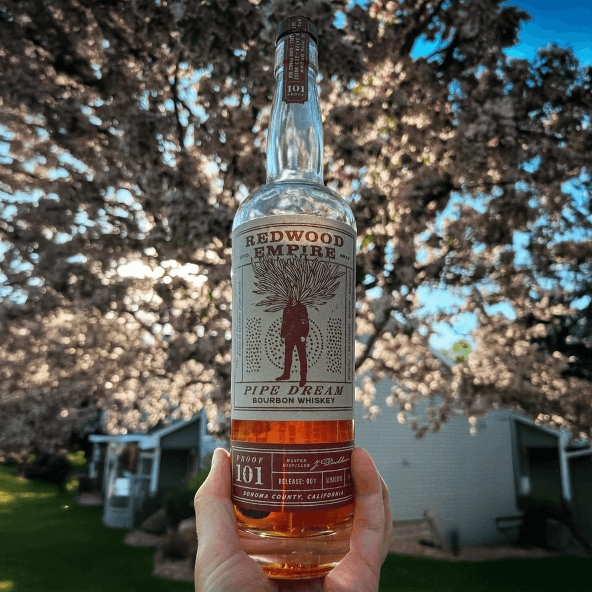 Redwood Empire Pipe Dream 101 Proof bourbon whiskey bottle against blooming trees.