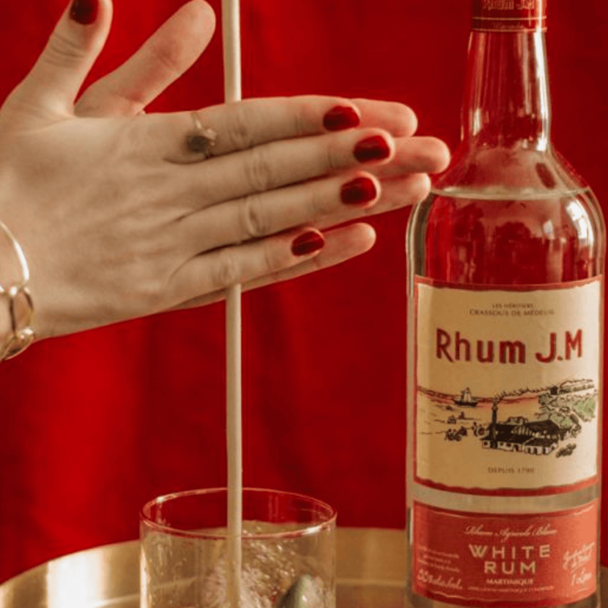 Rhum J.M Agricole Blanc 110 Proof bottle with cocktail on a table, showcasing vibrant red background.