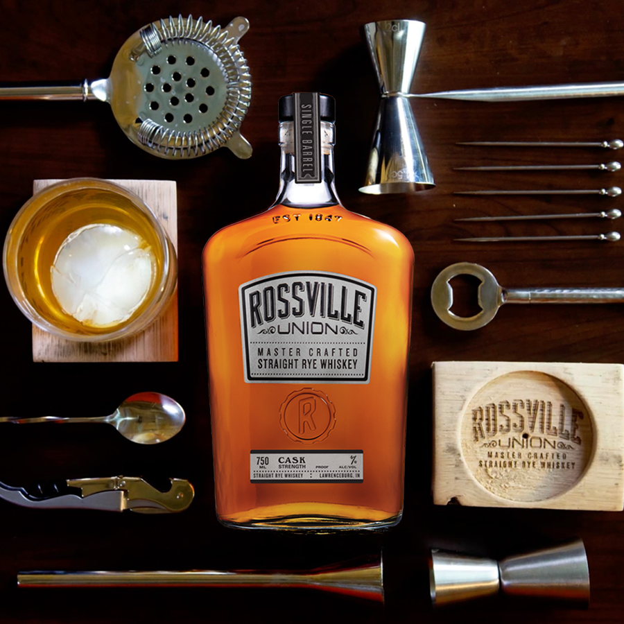 Rossville Union Whiskey bottle surrounded by cocktail tools and a glass of whiskey on a wooden table.