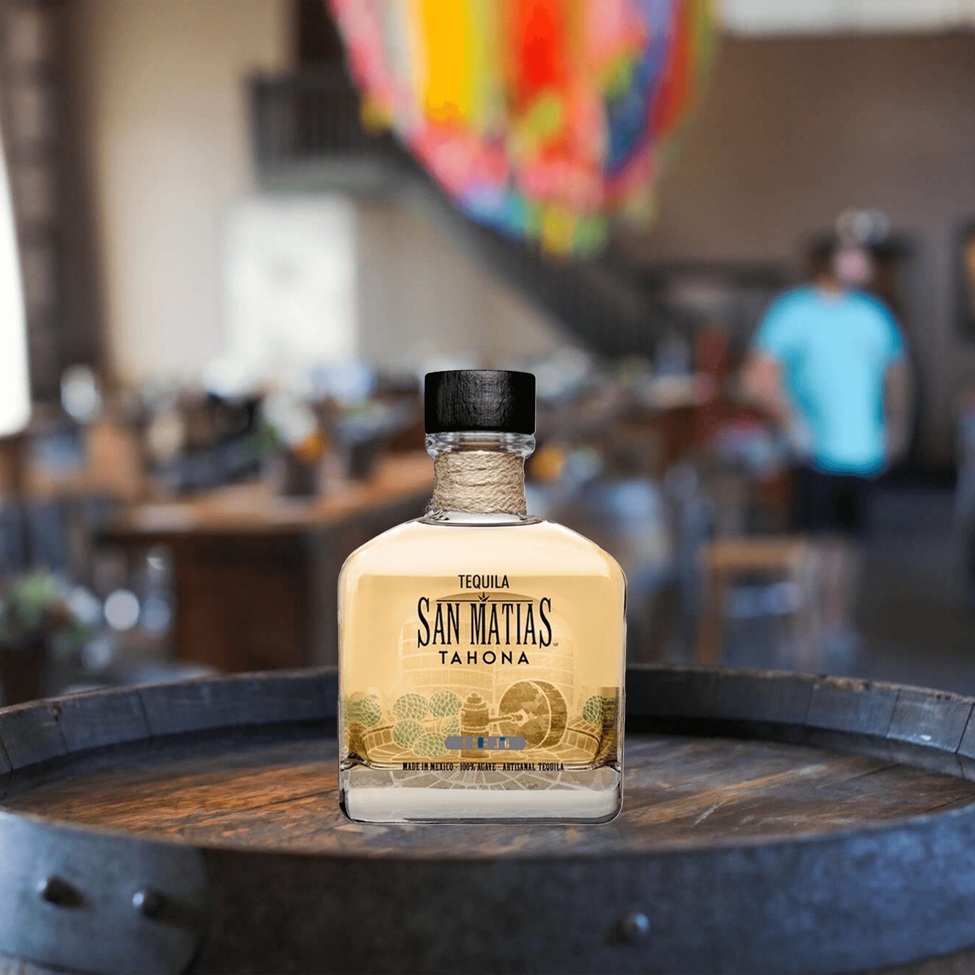 San Matías Tahona Añejo Tequila bottle on a wooden barrel in a vibrant setting, showcasing elegance and tradition.