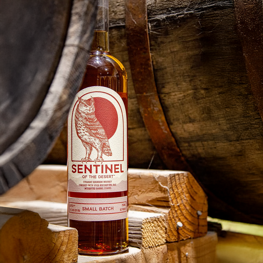 Bottle of Sentinel Bourbon of the Desert displayed with oak barrels in the background.