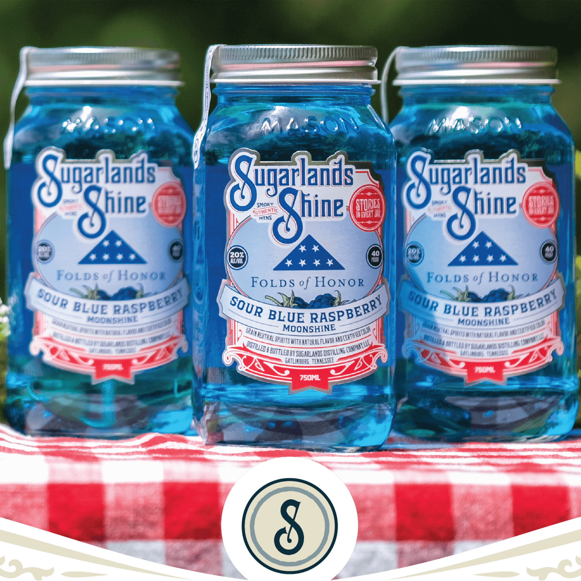 Three jars of Sugarlands Shine Folds of Honor Sour Blue Raspberry Moonshine on a checkered tablecloth.