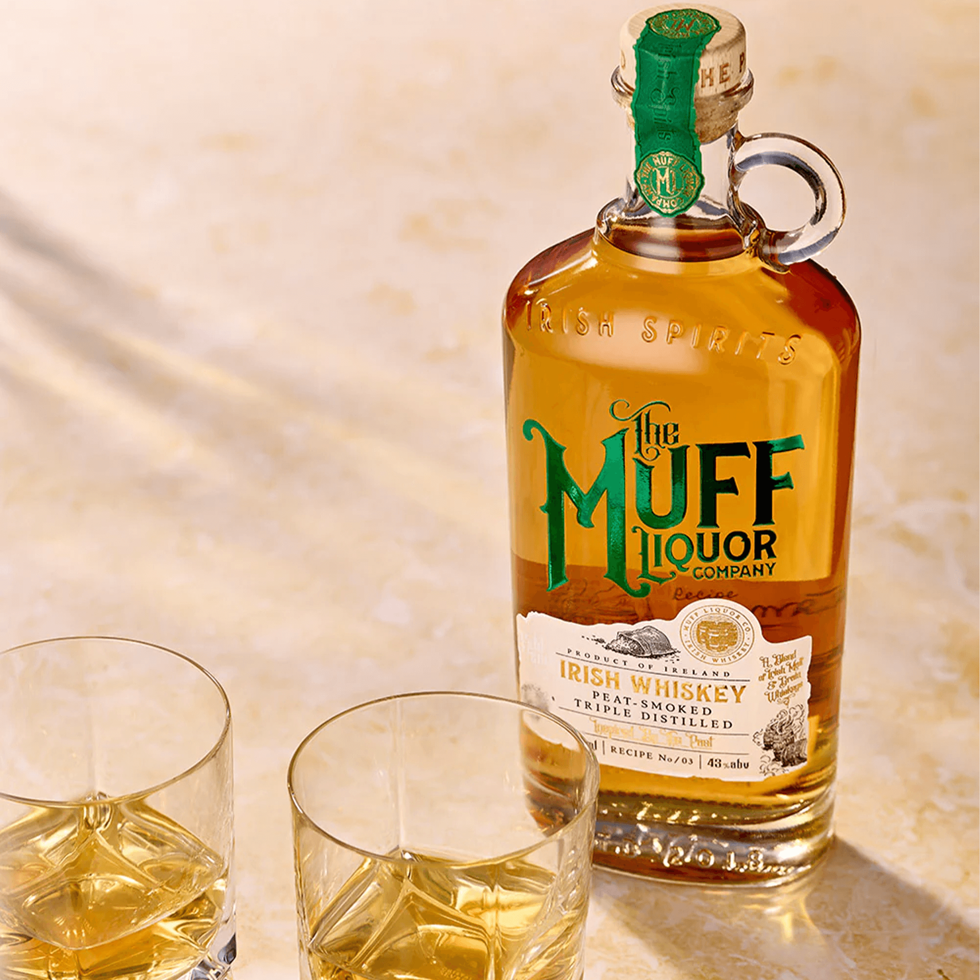 Bottle of The Muff Liquor Peat Smoked Irish Whiskey with two glasses on a wooden surface.