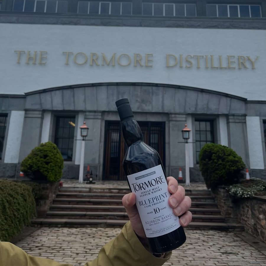Hand holding Tormore Blueprint 10 Year Bourbon in front of The Tormore Distillery, showcasing Speyside whisky.
