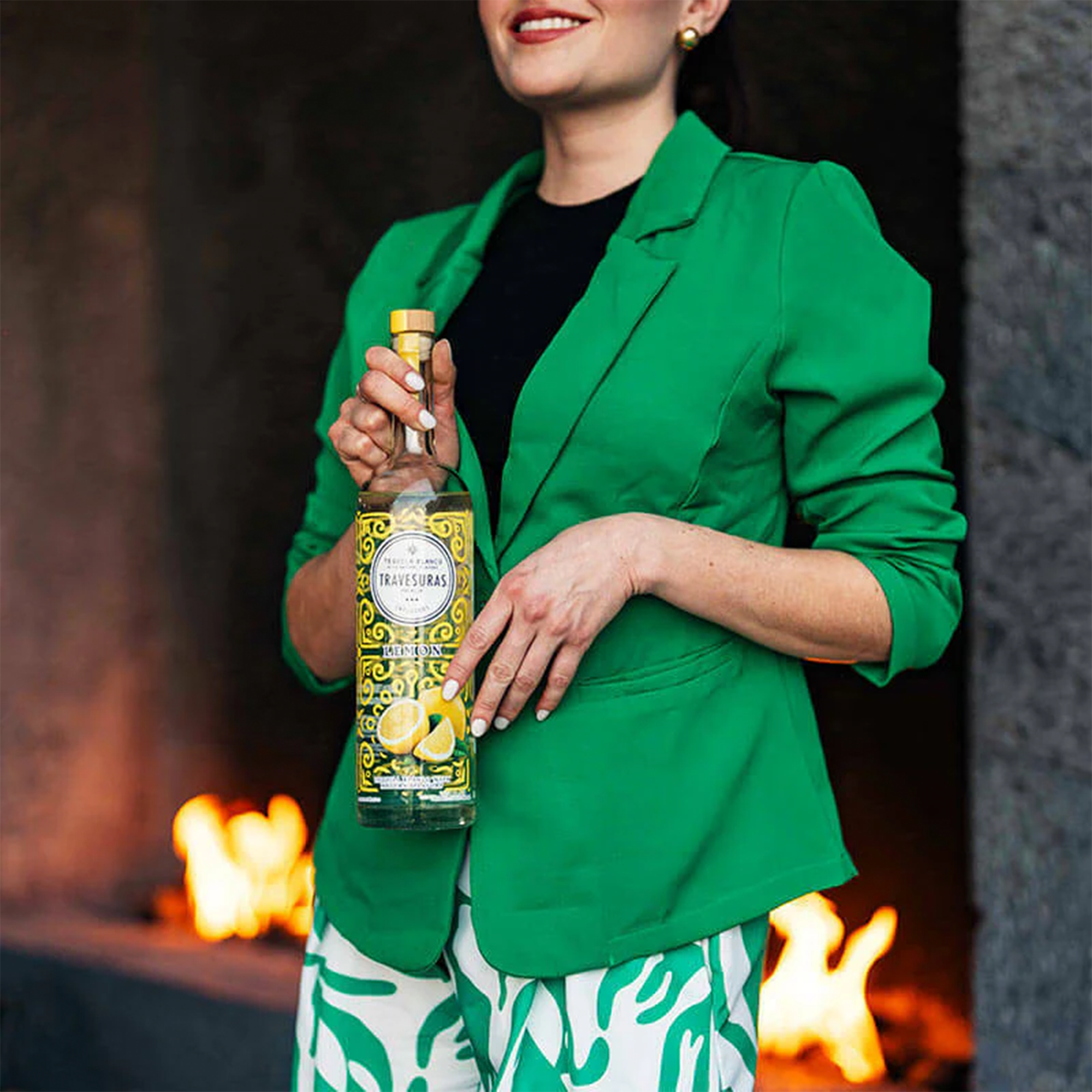 Person in a green blazer holding a bottle of Travesuras Lemon Tequila in front of a fireplace.