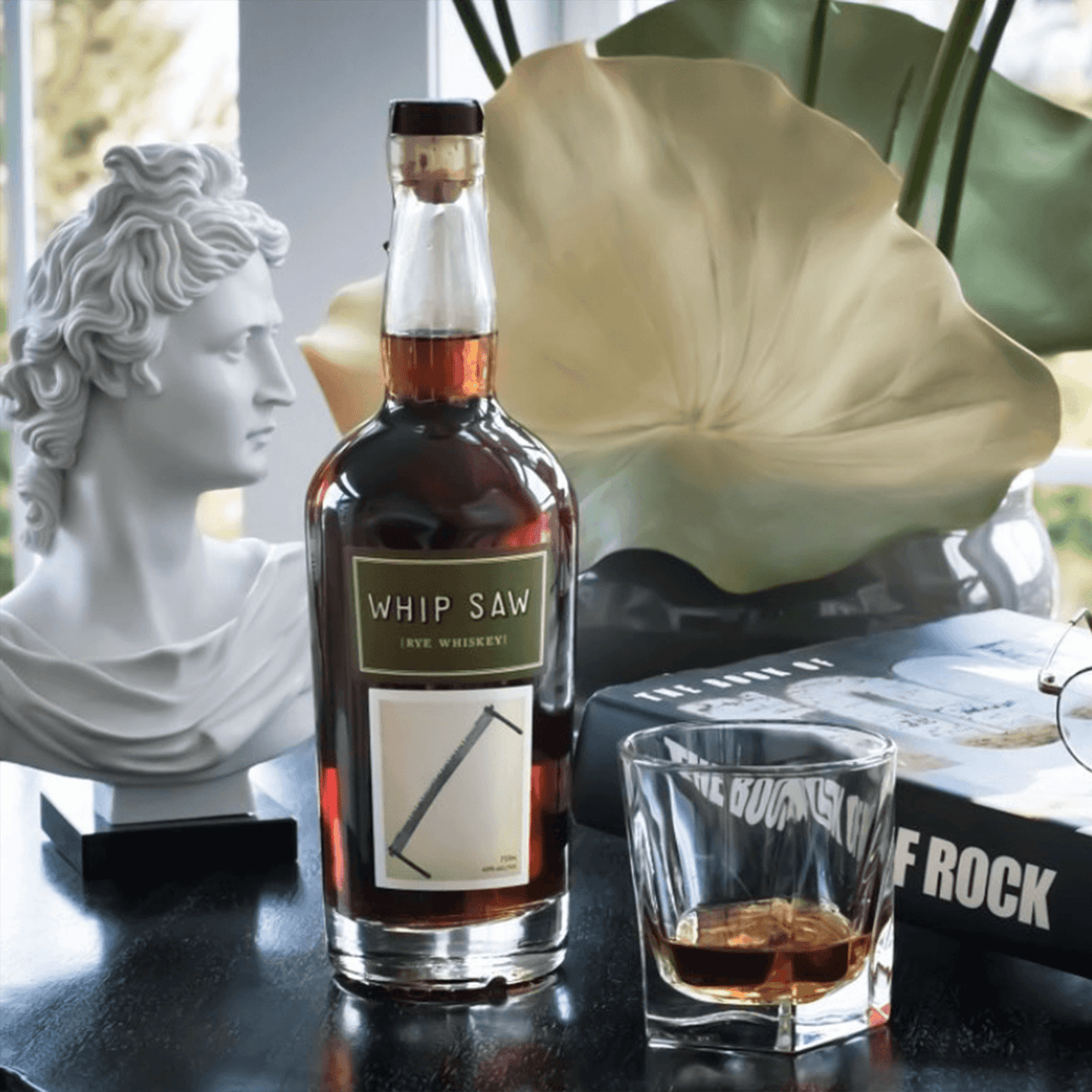 Whip Saw Rye Whiskey bottle beside a glass, sculpture, and books in a stylish setting.
