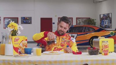 Man in Eggo jersey pours syrup over waffles at a festive breakfast table with cars in the background.