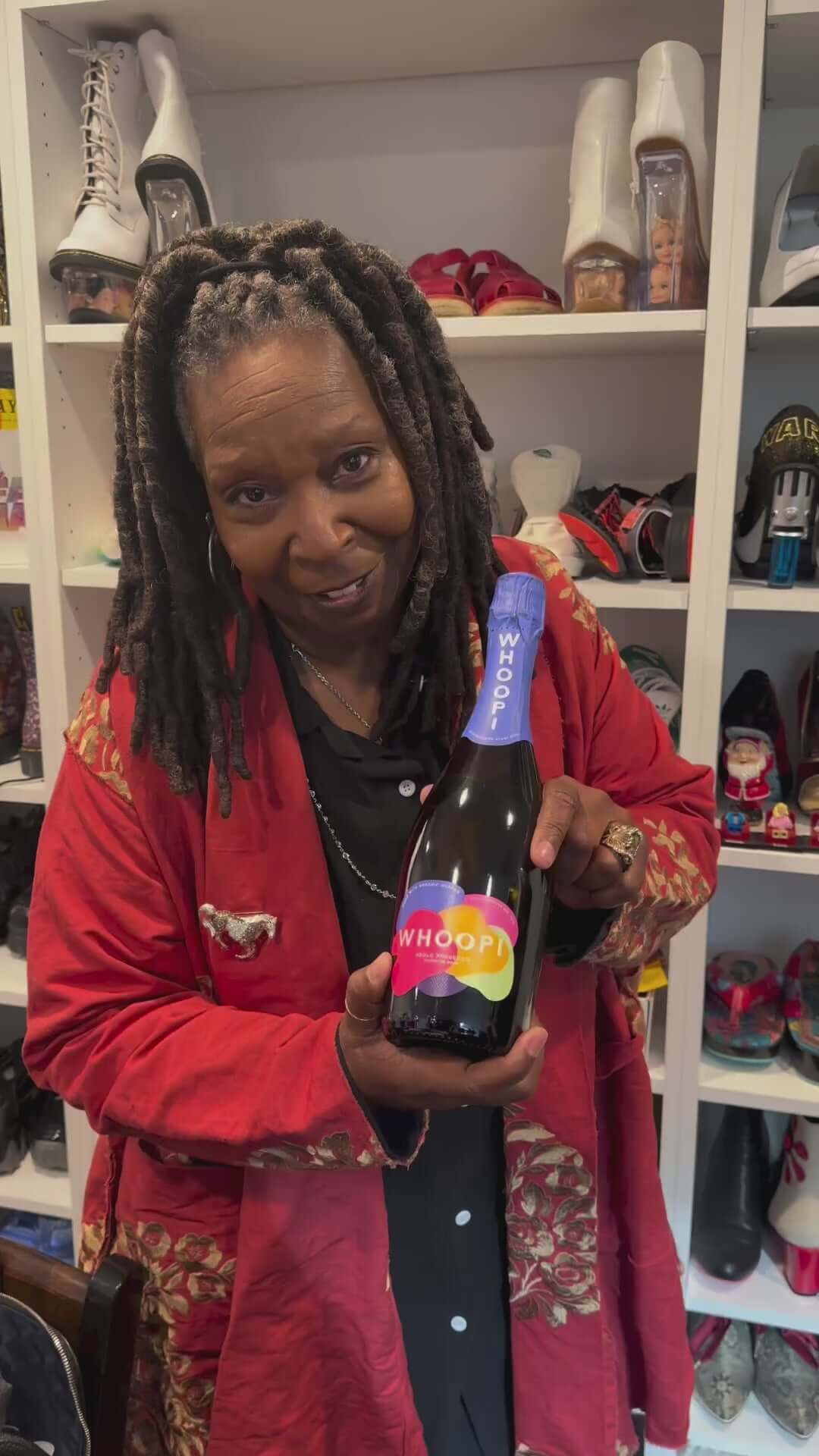 Whoopi Goldberg proudly holds a bottle of Whoopi Prosecco with a vibrant label in a stylish setting.