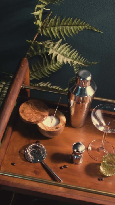 Cocktail preparation setup with a shaker, strainer, bowls, and foliage on a wooden tray.